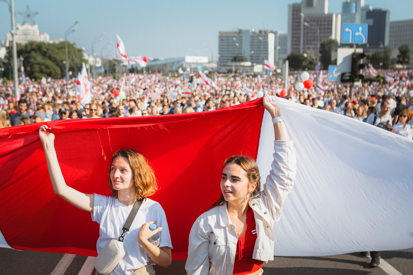 Två belarusiska kvinnor står håller upp landets flagg, i bakgrunden ser man ett folkhav av protesterande belaruser