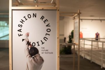 Ett foto av den uppbyggda utställningen Fashion revolution - the future of textiles. I en trämodul visas en bild på utställningen logga och ett modefoto med en kvinna bakifrån som sträcker upp armarna. I bakgrunden syns resten av utställningen oskarp.