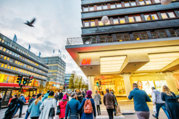 A crowd walking on a shopping street passing an H&M store. The sky is grey and a pigeon is flying right across.