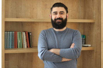 A photo showing a man with dark hair and beard, smiling standing in front of a bookshelf.