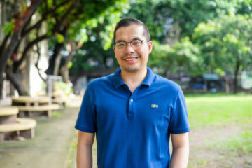 A man wearing glasses, a blue shirt, smiling, standing outside with trees in the background. 