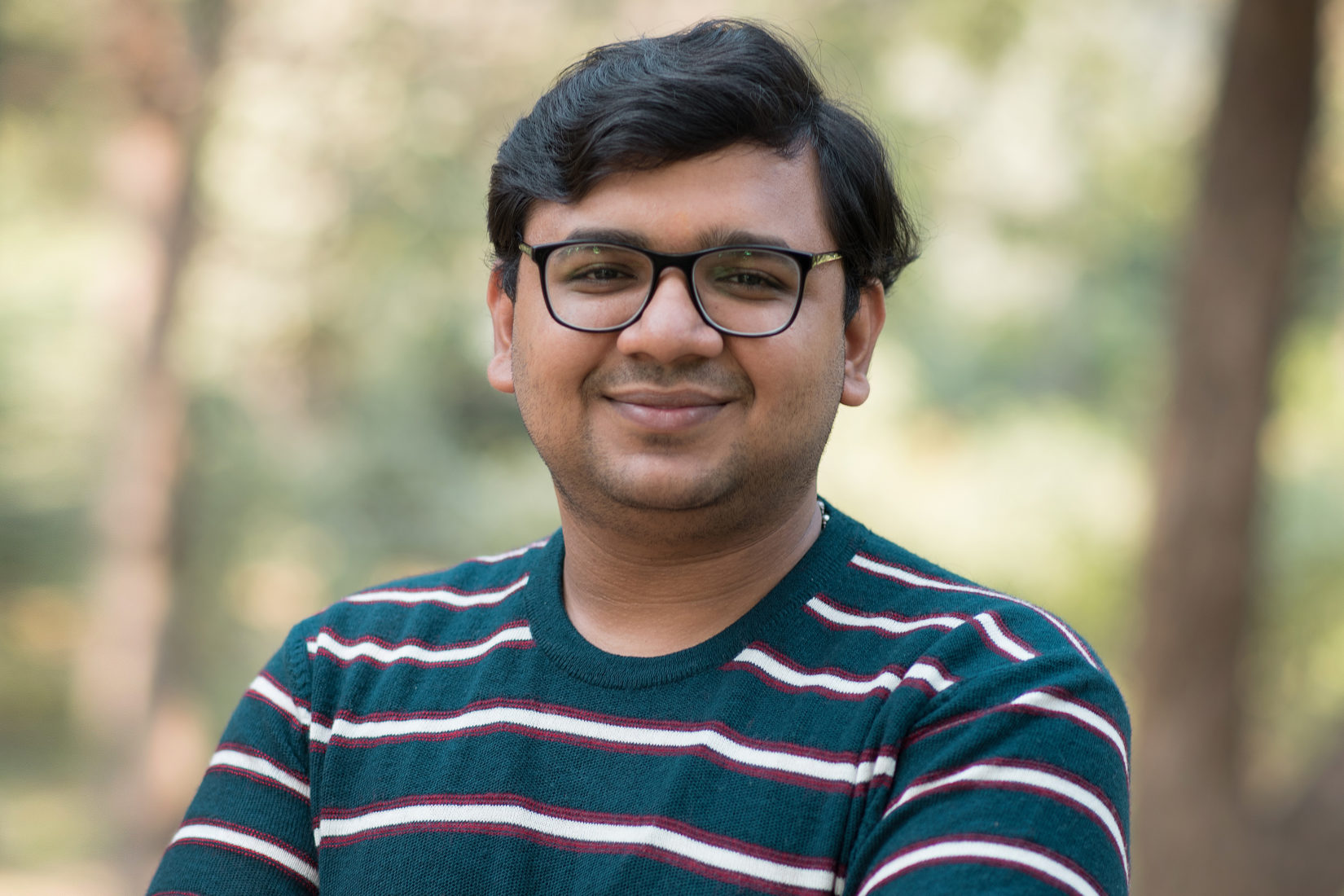 A man wearing glasses and a striped sweater, smiling, standing outside. 