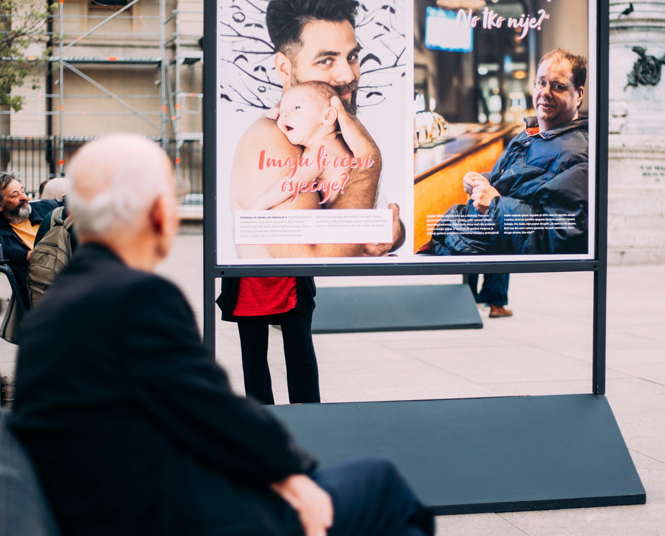 An old man sits in a open space, a square ,and looks at an image showing a bare-breasted dad holding a naked infantnext to him. 