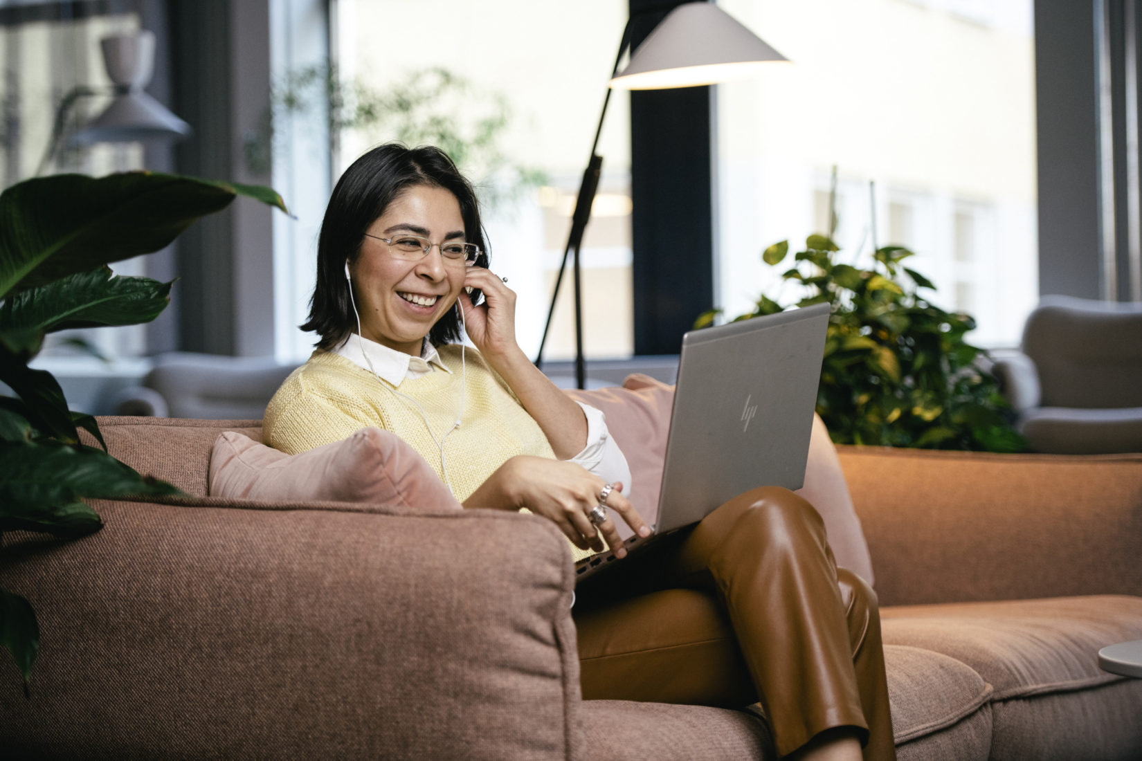 margareta_bloom_sandebäck-conncted_sweden-8169 En glad kvinna sitter i en rosafärgadsoffa och pratar i telefon med en uppfäld laptop i knät.