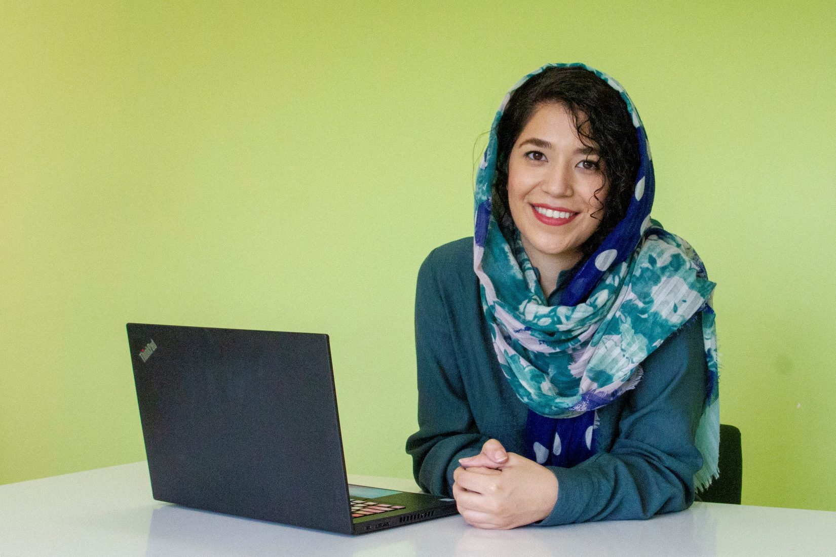 A woman sitting in front of a laptop, wearing a green jacket and a shawl aorund her hair.