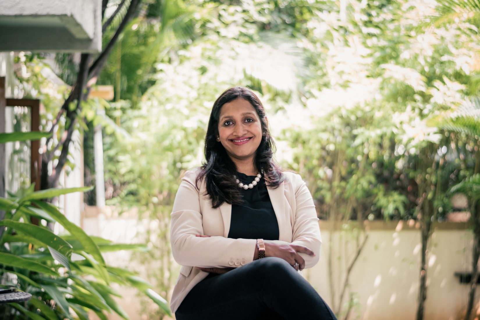 A woman smilin, with dar long hair, sitting with her arms crossed, wearing a white suit. In the background green trees. 