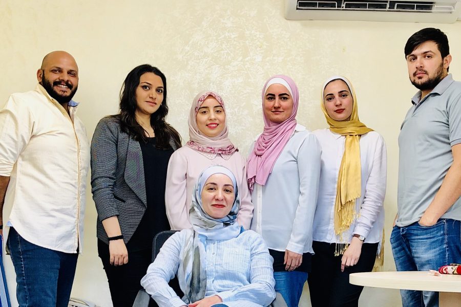 A picture of seven people. Five women and two men. One woman is sitting on a chair in front of the rest of the people who are standing in a line behind her. They are smiling .