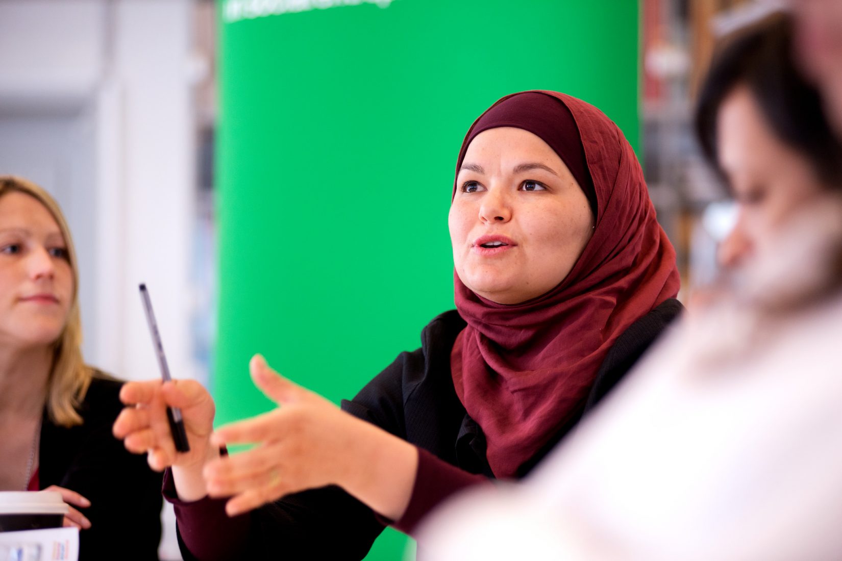A woman in a red/brown hijab. She is standing infront of a green wall and is having a discussion with people outside of the picture.