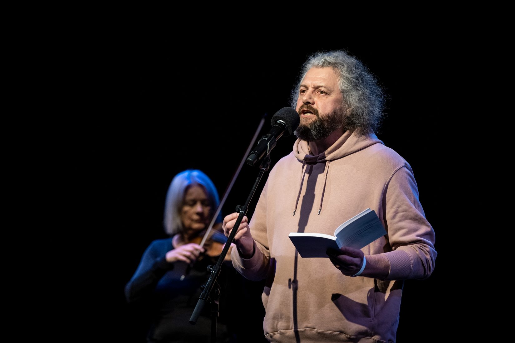Den belarusiske poeten Dmitrij Strotsev står på en teaterscen med en bok i handen och reciterar en dikt, i bakgruden ser man en kvinnlig violinist.