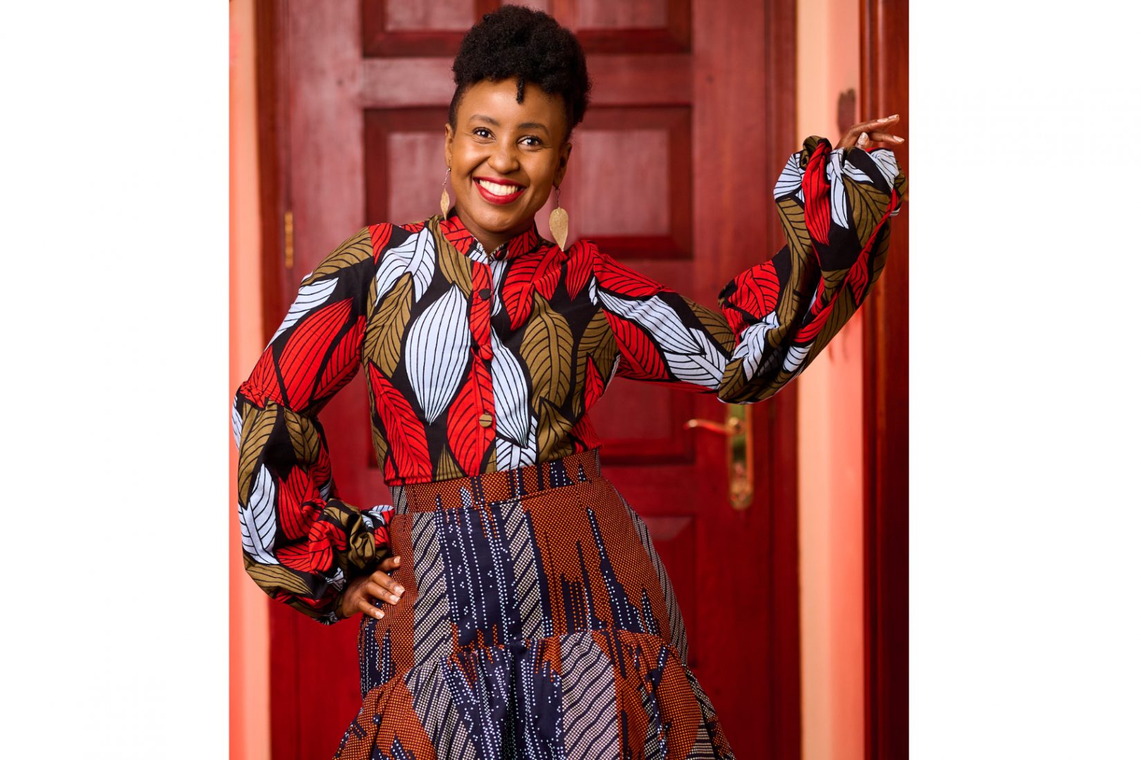 Woman smiling, wearing patterned shirt and skirt standing in front of a door.