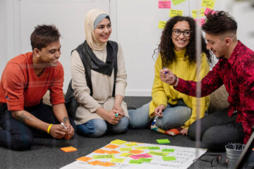 Four young people sitting in a workshop on the floor. In front of them they have colourful post-its. They are happy and in a discussion.