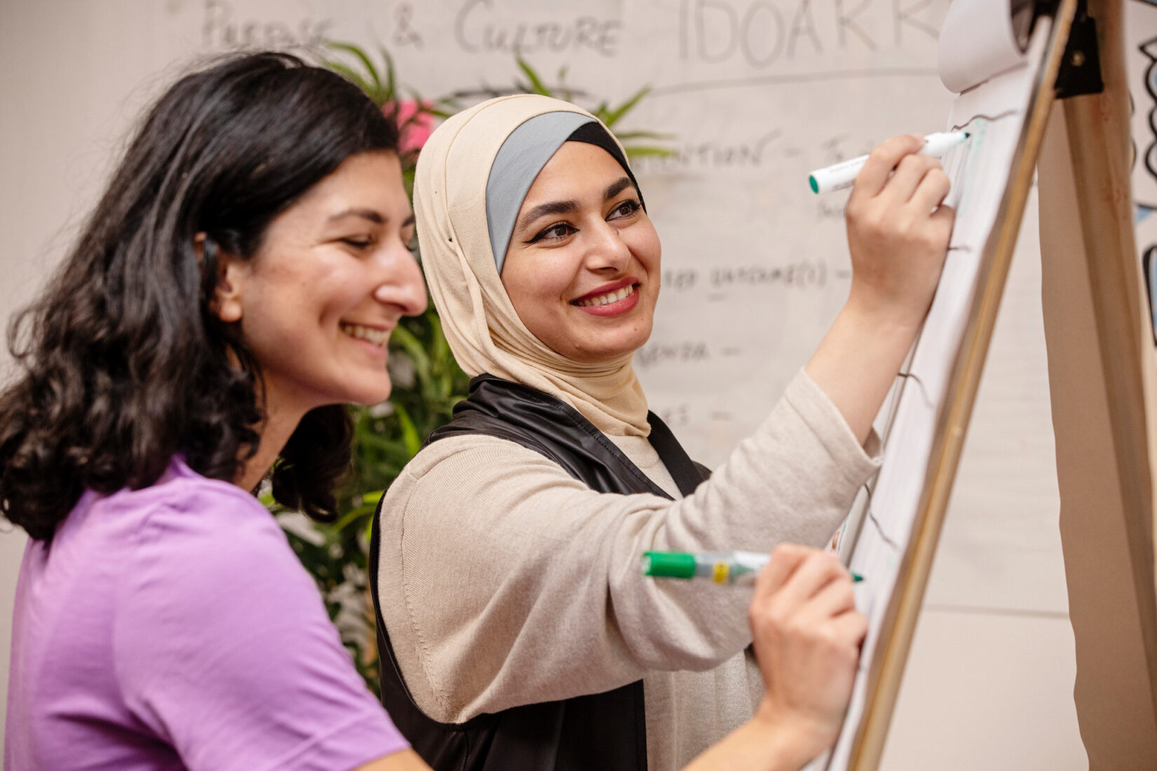 Two women writing on a whiteboard. The one in the front has dark hair and a purple t-shirt. The one in the background wears hijab. Both look happy and seem to enjoy their company..