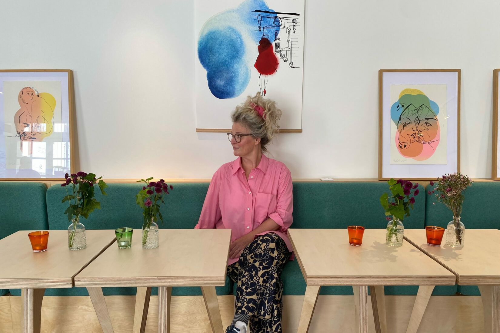 Stina sitter vid ett bord i kafét. Bakom henne hänger en av hennes konstverk.