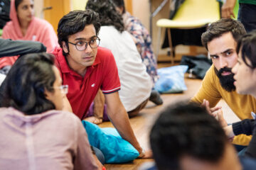 A young man in a red t-shirt, with dark hair and glasses is sitting on the floor listening engaging in a discussion with peers.