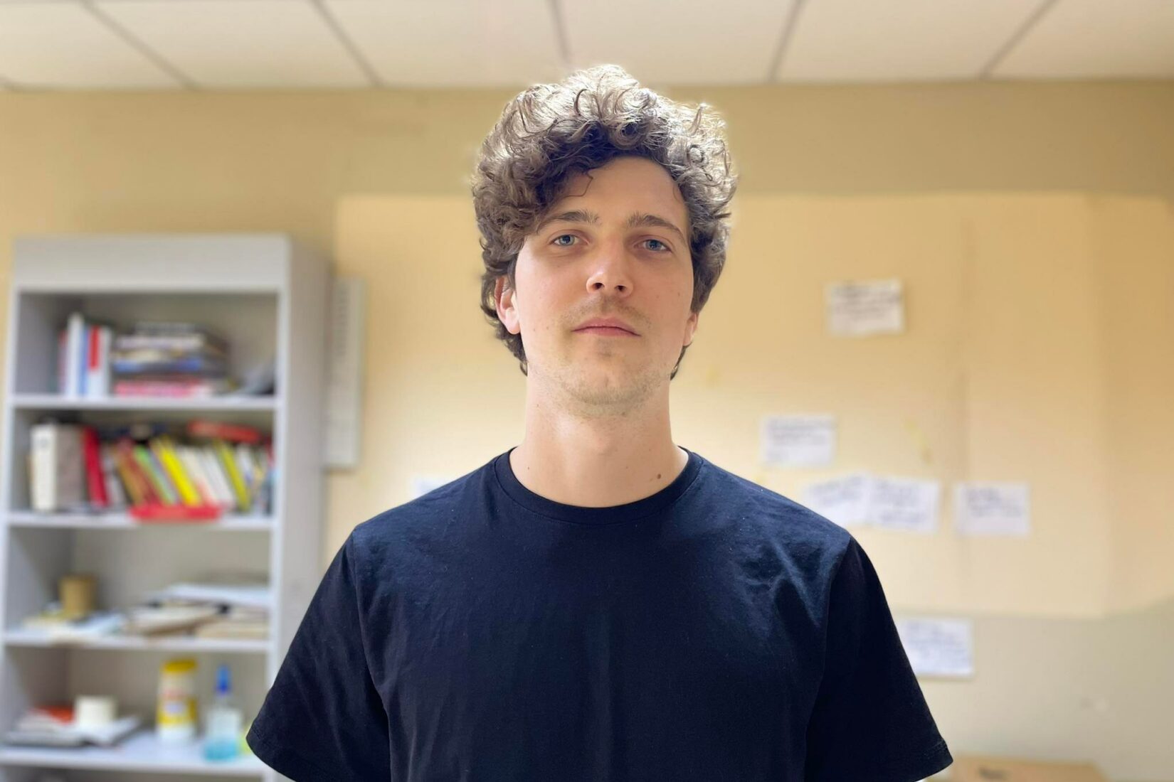 Portrait of a young man standing in classroom. Wears a black t-shirt.