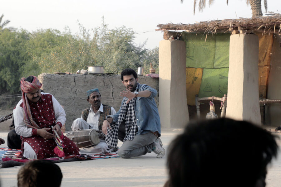 Three men sitting on the ground. Two of them are holding traditional Pakistani instruments in their hands. 