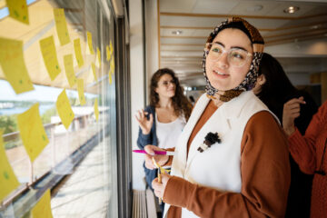 A woman is standing in front of a window with many post-its on it. She is wearing a weil and glases.