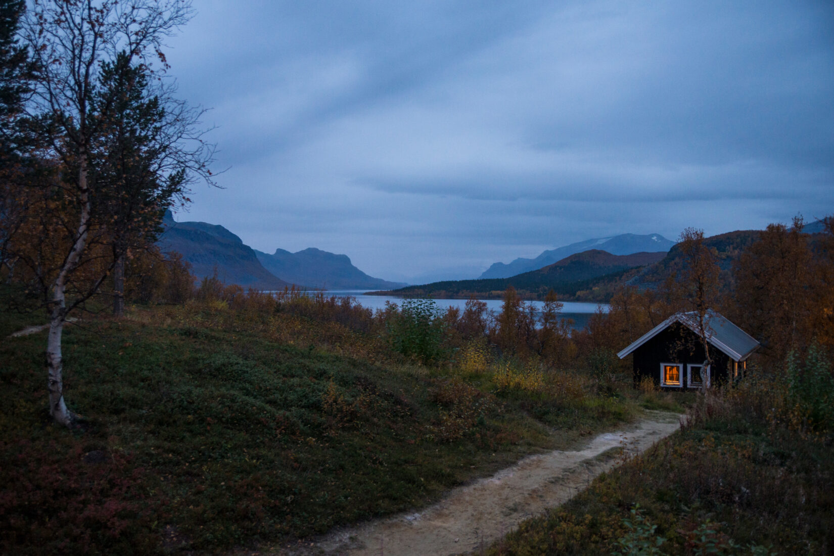 Liten röd stuga vid sjö och skog i skymning.