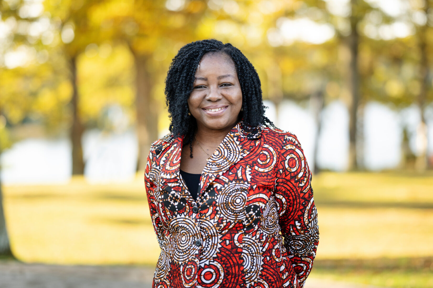 Woman in a red/white/black blouse staning outdoors smiles to the camera.