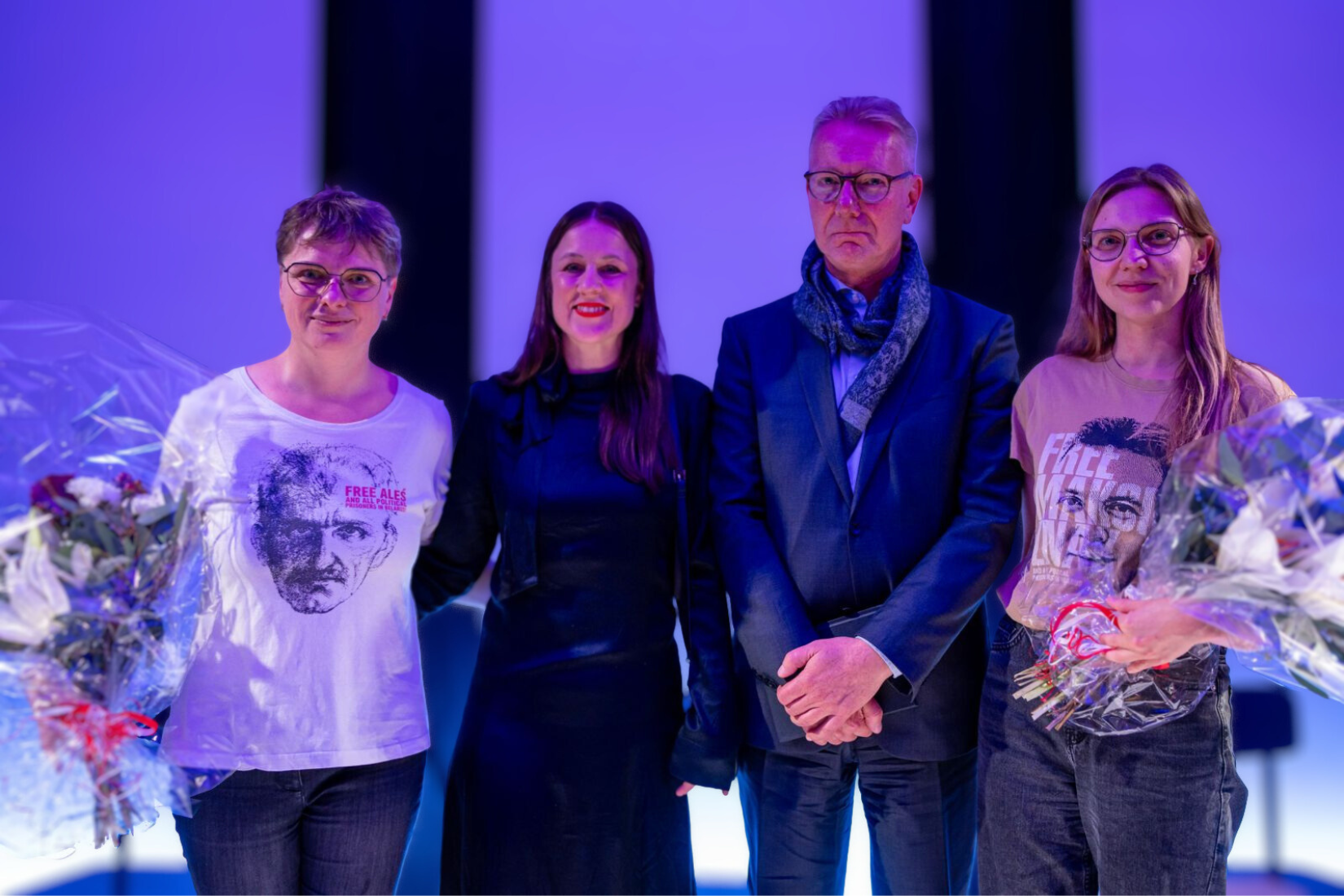 Three women and a man are smiling and standing together on a stage.