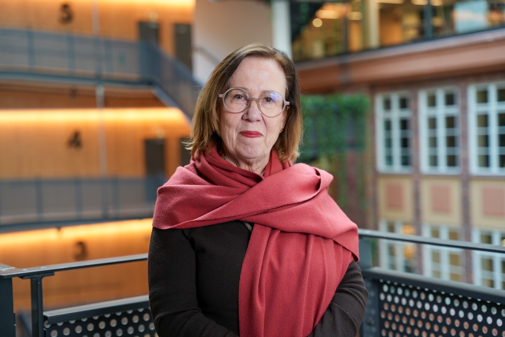 Person står inomhus vid ett räcke, iklädd mörk tröja och en stor rosa sjal. Bakgrunden visar en modern byggnad med glaspartier, metallräcken och träpaneler samt stora fönster med rutmönster